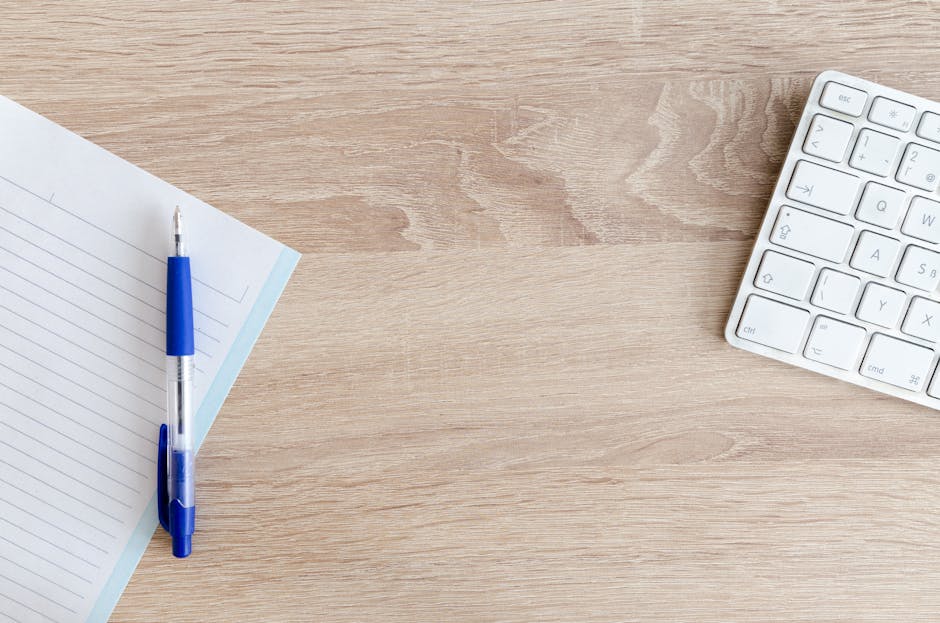 A minimalist workspace with a keyboard, notepad, and pen on a wooden desk.