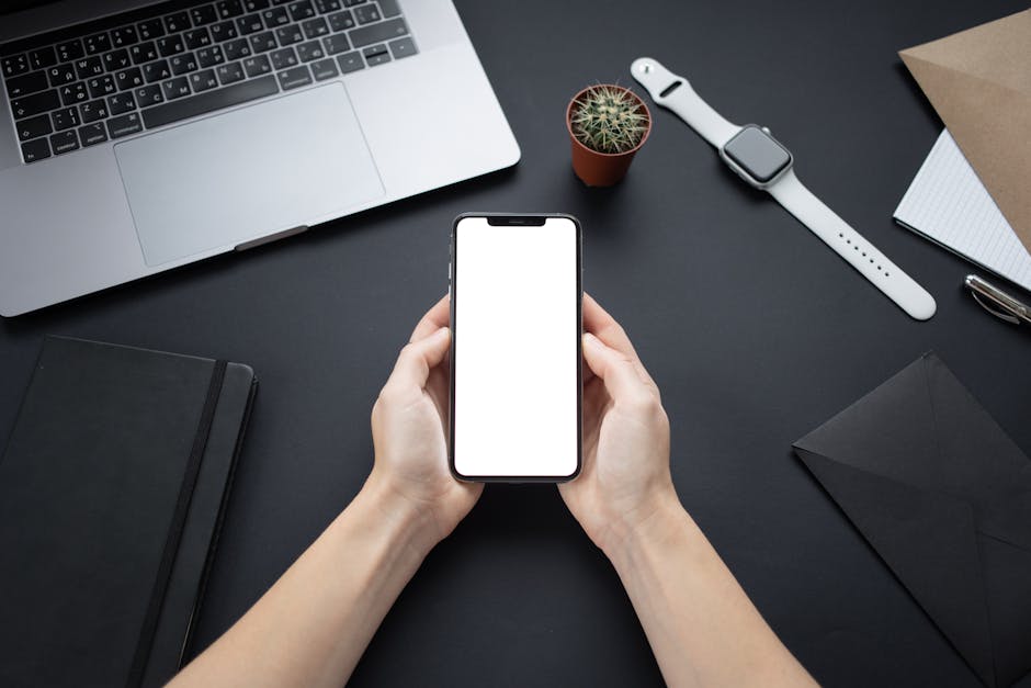 Hands holding a smartphone with blank screen on a minimalist office desk setup.