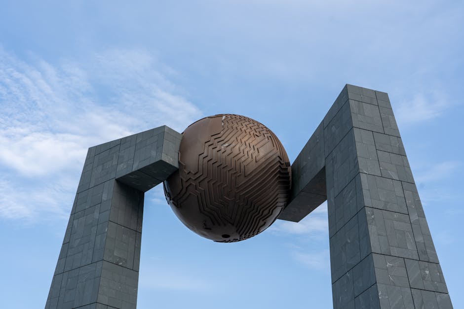 A unique modern sculpture with a large metal sphere supported by two stone pillars set against a clear blue sky.