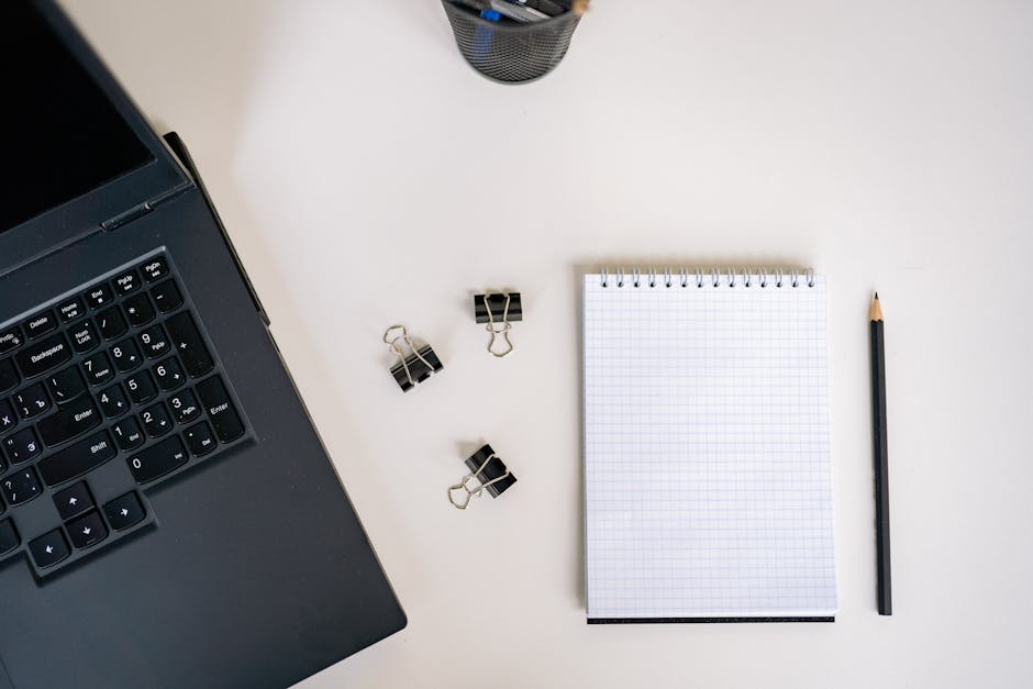 Top view of an office workspace featuring a laptop, notepad, pencil, and binder clips on a clean desk.